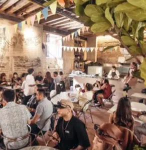A group of people sit at tables in a decorated café in Bogotá, featuring exposed brick walls, bunting, and a visible kitchen area.